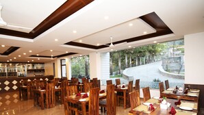 A restaurant with wooden tables and chairs, a bar area, and large windows offering a view of a landscaped area. - Ashoka Resort Pvt. Ltd (Kabilas)