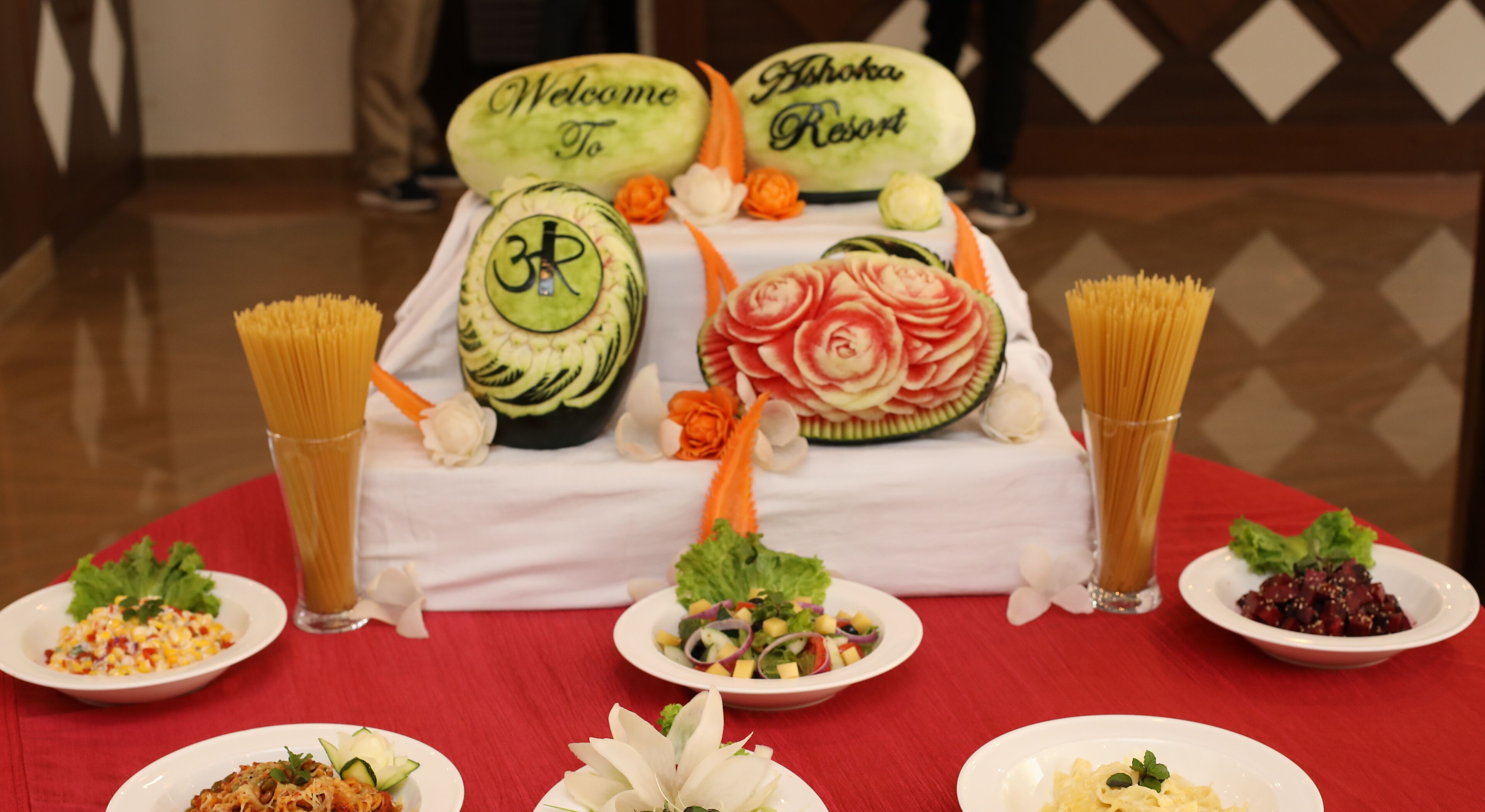 A table with various dishes, including a watermelon carved into a rose, pasta, and salads, set on a red tablecloth.