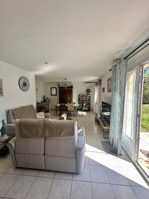 A living room with a sofa, dining table, and chairs, a bookshelf, and a large window with curtains.