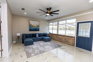 A living room with a sectional sofa, a ceiling fan, a large window, and a door. - Bayou, Blues & Views (Pensacola)