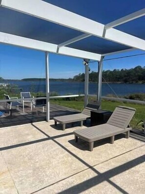 Une terrasse extérieure avec des chaises longues et un espace abrité, offrant une vue sur un plan d’eau et la verdure.