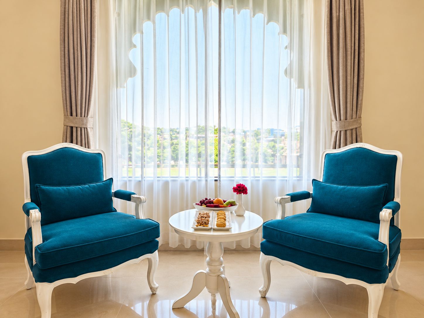 A room with two blue upholstered chairs, a white table with a fruit bowl and flowers, and a view through sheer curtains.