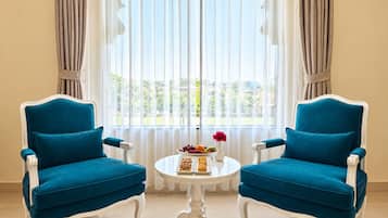 A room with two blue upholstered chairs, a white table with a fruit bowl and flowers, and a view through sheer curtains.