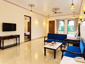 A hotel room with a TV, a sofa, a coffee table with fruit, a telephone, and a balcony with a view. - Trulyy The Shakti Vilas (Udaipur)