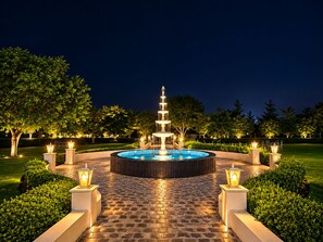 A nighttime outdoor fountain with a circular stone pathway, surrounded by evenly spaced lanterns and trimmed hedges.