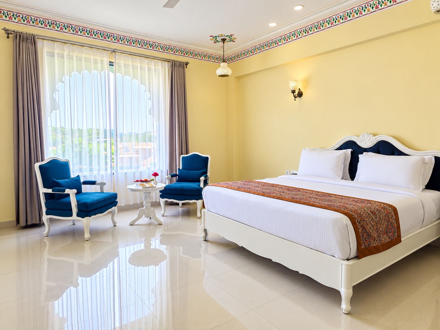 A hotel room with a large bed, two blue armchairs, a small table, and a window with curtains.