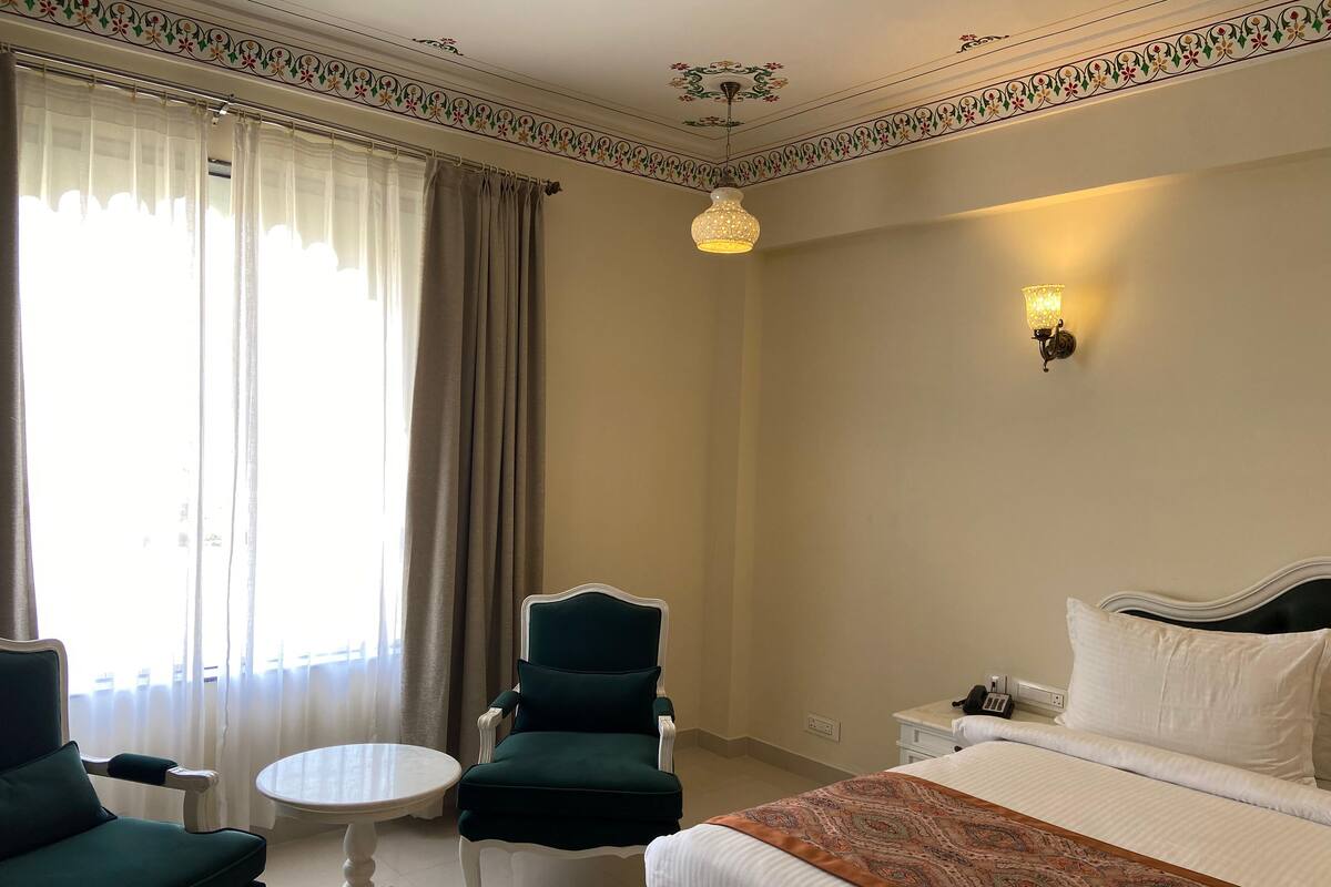 A hotel room with a bed, two chairs, a small table, a decorative ceiling, and a wall-mounted lamp.
