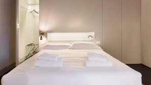 Un lit bien fait, avec du linge de lit et des serviettes blanches, une lampe de chevet et une vue sur une salle de bain équipée d’un porte-serviettes.