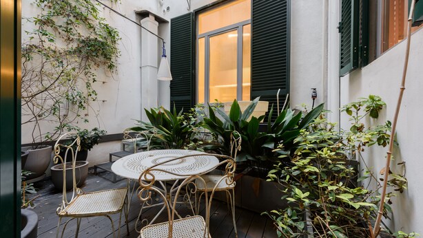 Une terrasse avec une table ronde et quatre chaises, entourée de plantes en pot et d’une fenêtre aux volets verts.