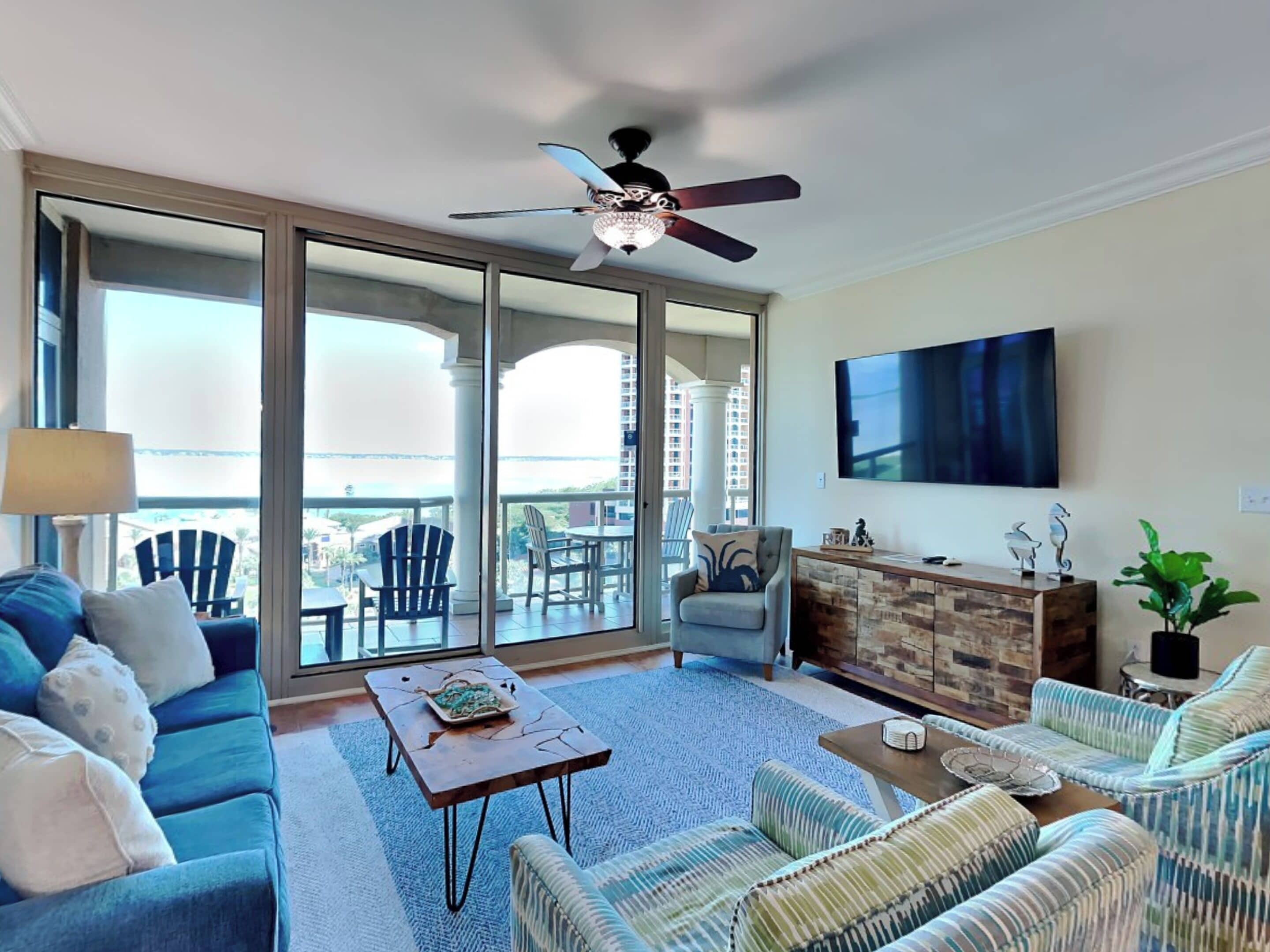 A spacious living room with a sofa, armchairs, a coffee table, and a TV. There is a balcony with chairs and a view of buildings.