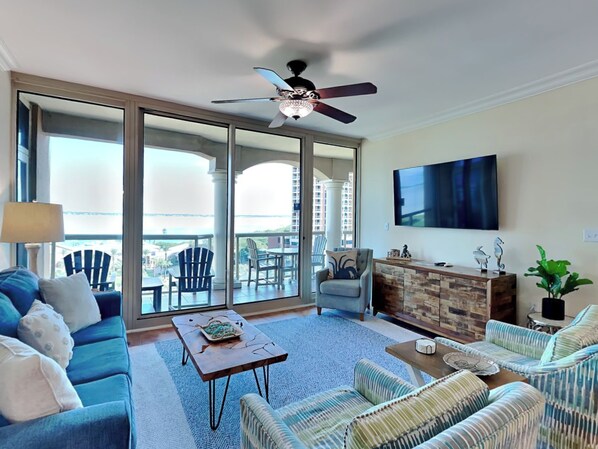 A spacious living room with a sofa, armchairs, a coffee table, and a TV. There is a balcony with chairs and a view of buildings.