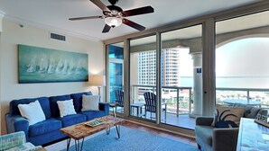 A spacious living room with a blue sofa, a wooden coffee table, and a large window offering a view of the city skyline and ocean.