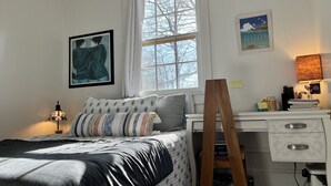 Ein Schlafzimmer mit Bett, Schreibtisch, Lampe, einer Leiter und einem Fenster mit Blick auf Bäume.