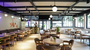 Un café espacioso con mesas redondas y sillas acolchadas, ventanas amplias y un diseño moderno de techo.