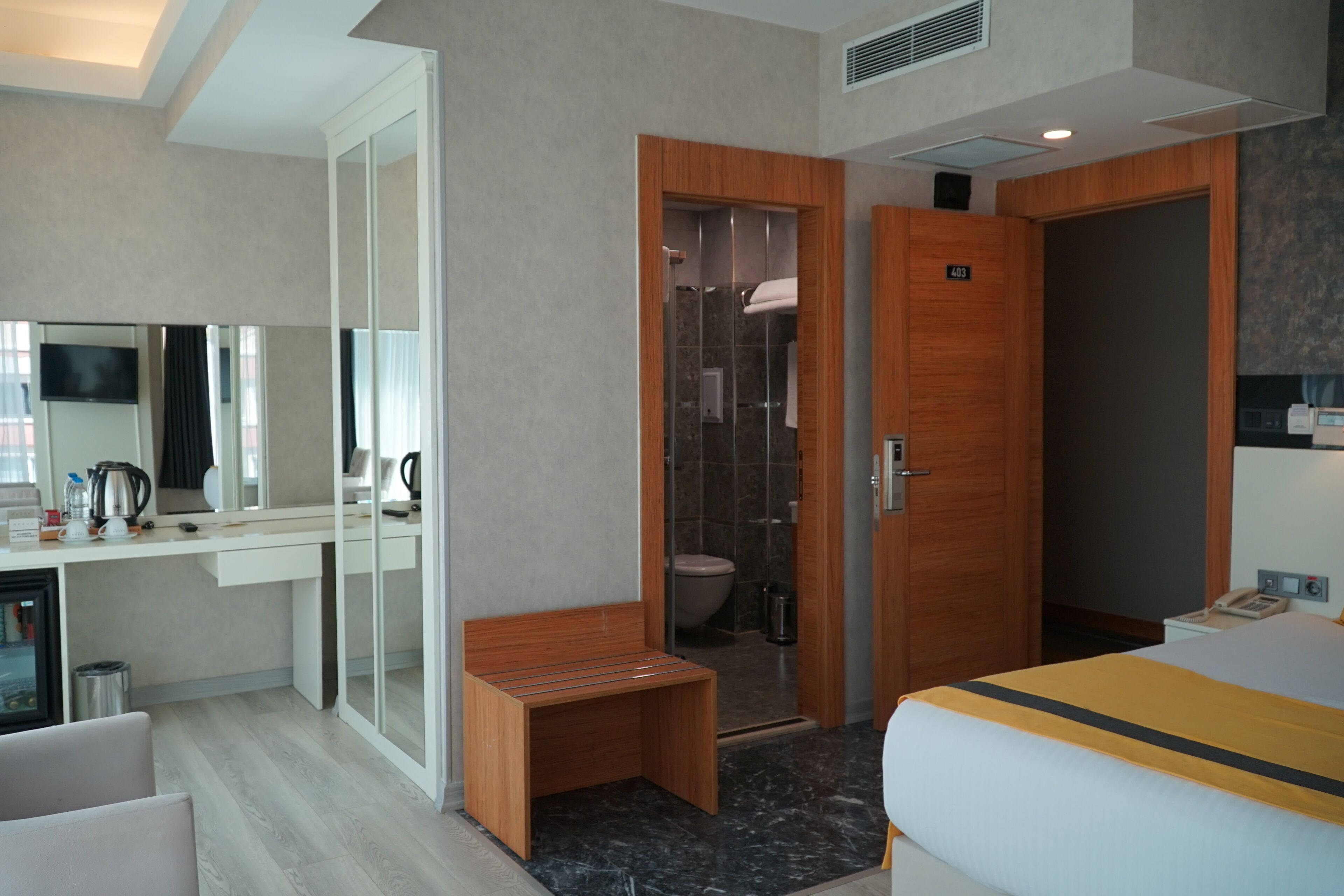 A hotel room with a bed, a wooden bench, a shower area, a toilet, a sink, and a mirror.