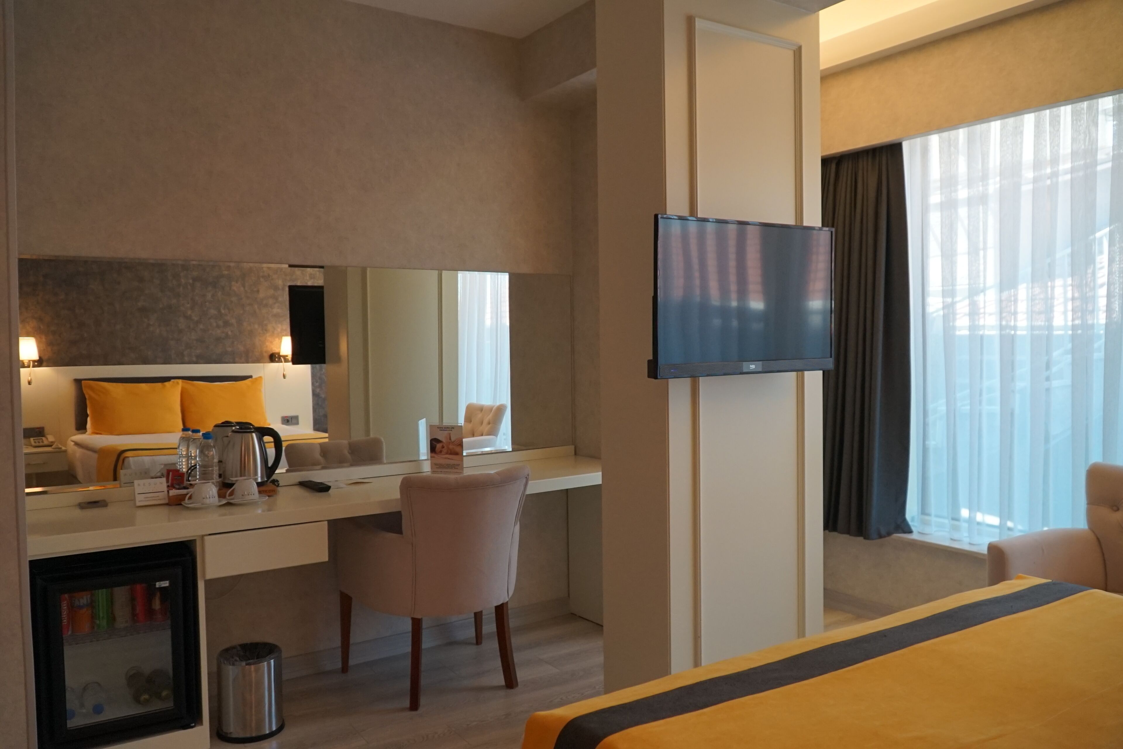 Habitación de hotel con cama, escritorio con silla, televisión, minibar y un ventanal grande.