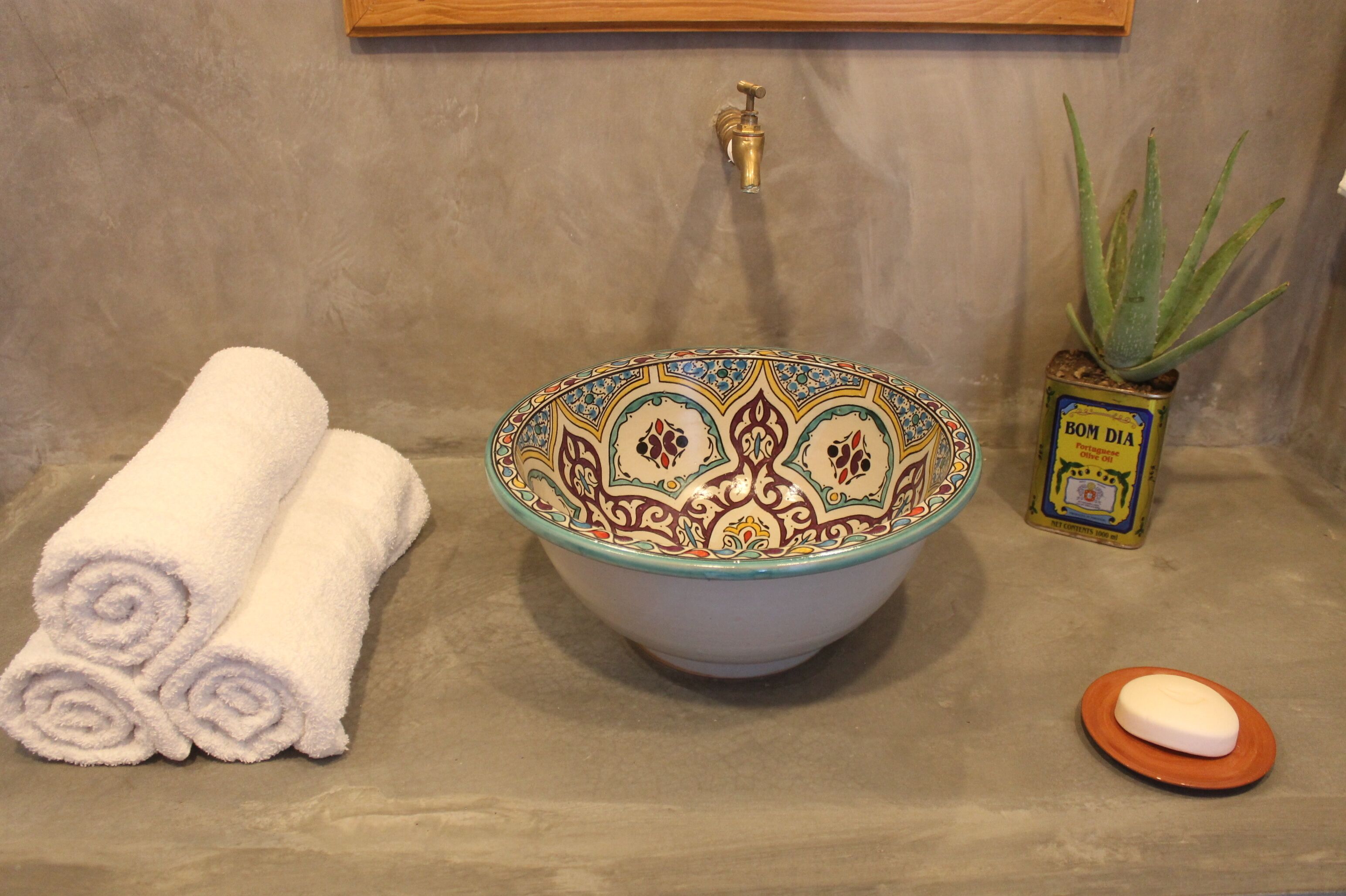 Un lavamanos de baño con un recipiente decorativo, una toalla enrollada, un jabonera y una planta de aloe vera en maceta.