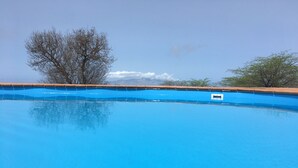 A clear blue swimming pool with wooden deck chairs in the foreground, surrounded by trees and mountains in the background. - La Fora Ecolodge (São Filipe)