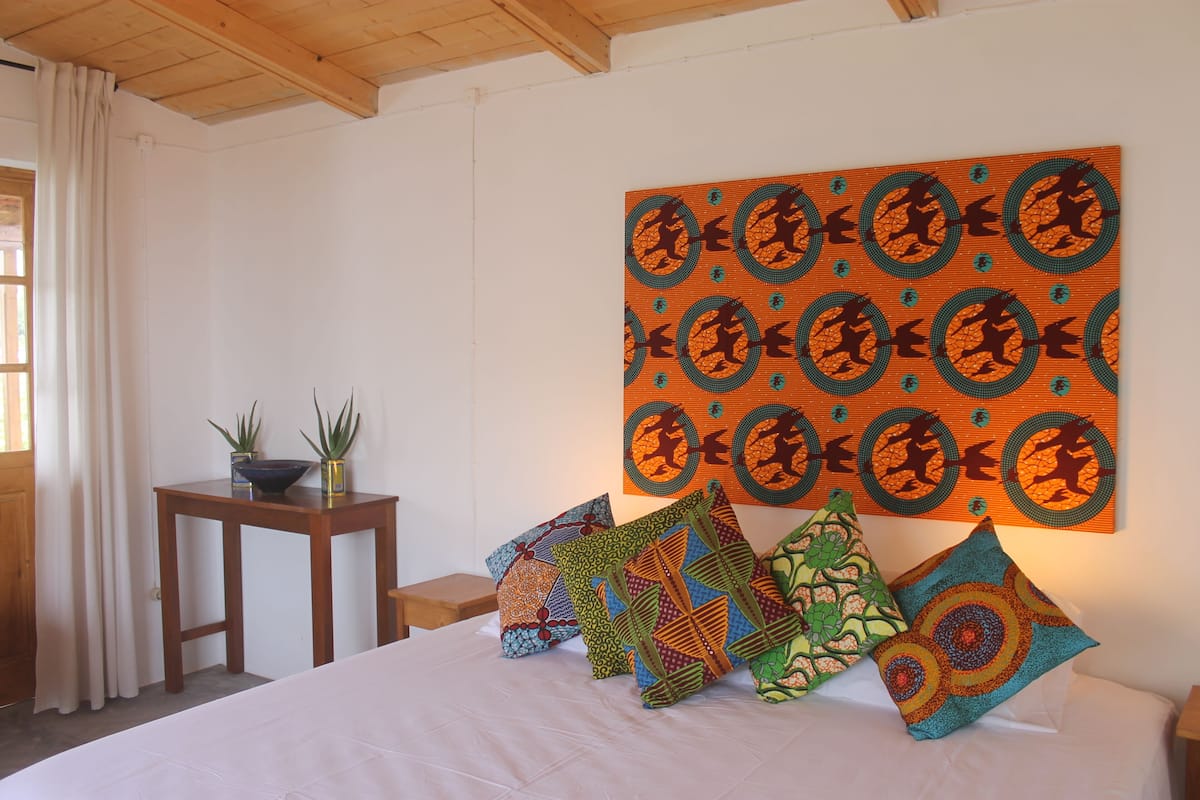 A bedroom with a large bed, multiple colorful pillows, a small wooden table with a plant, and a wall-mounted artwork.