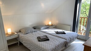 Ein Schlafzimmer mit zwei Betten, jeweils mit einem Nachttisch und einer Lampe. Ein Fenster bietet Blick auf Bäume.