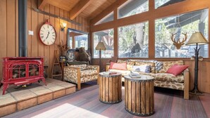 Une pièce chaleureuse avec un décor intérieur en bois, un poêle rouge, deux canapés à motifs et une grande fenêtre donnant sur les arbres.