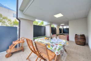 An outdoor dining area with a table set for two, wooden chairs, a fruit bowl, and a wine bottle.