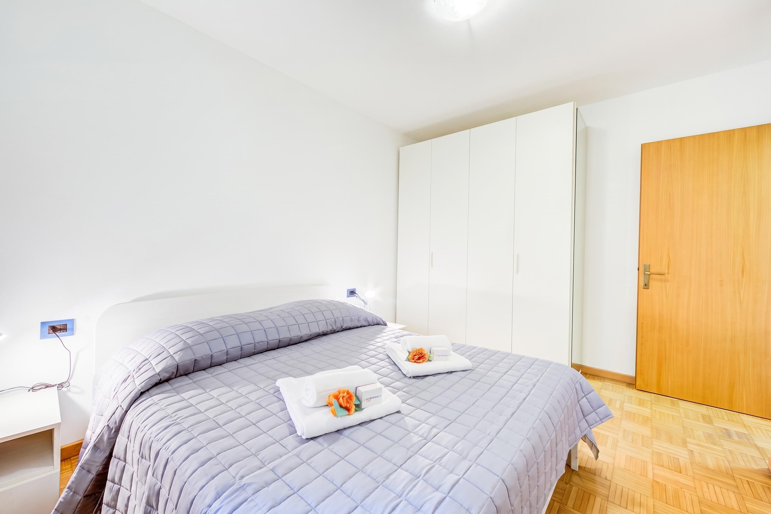 A bedroom with a bed, a nightstand, a wooden door, and a white wardrobe.