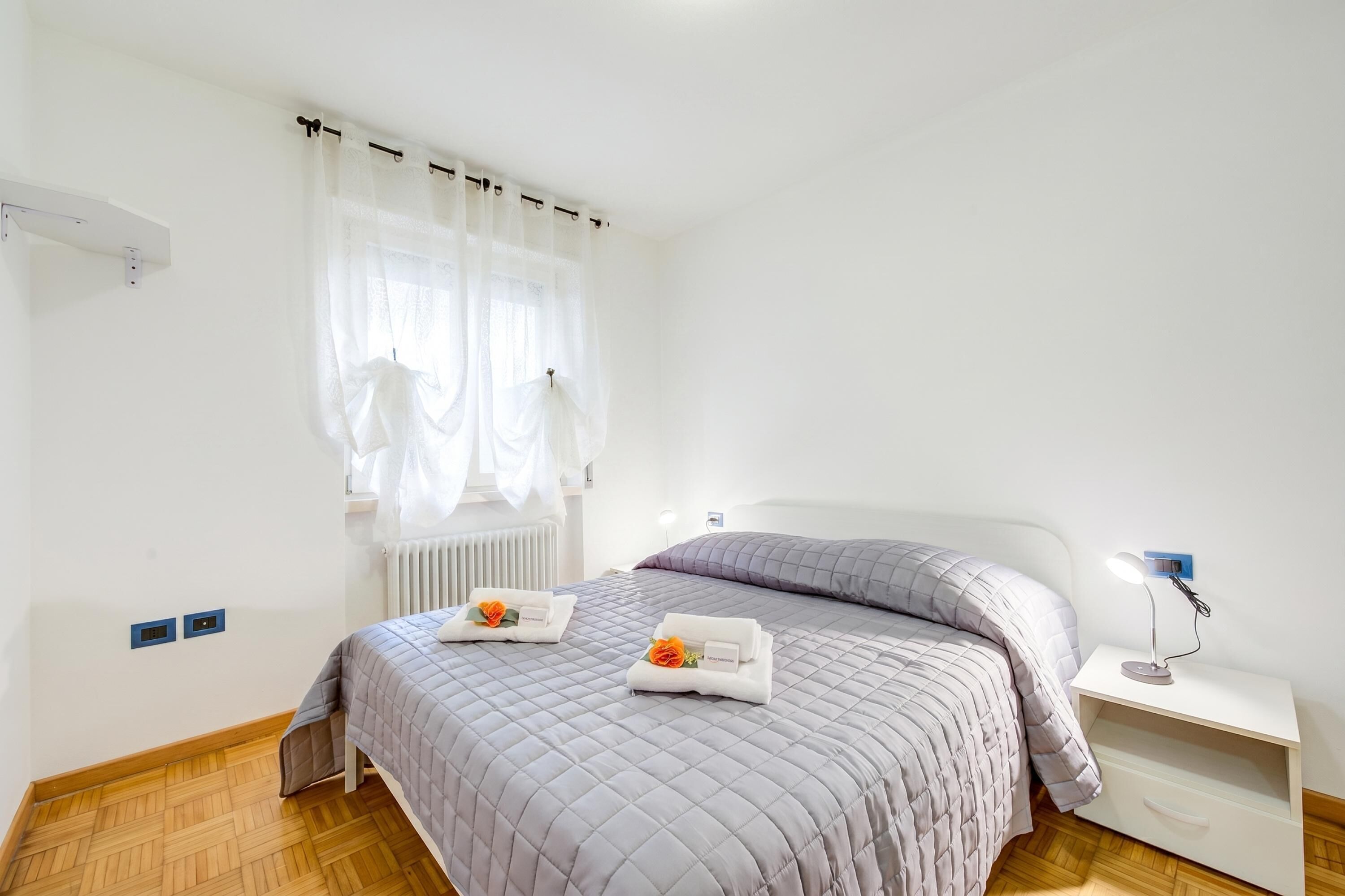 Un dormitorio con cama, mesita de noche, lámpara y ventana con cortinas.