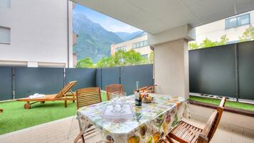 A patio with a table set for two, chairs, a bottle of champagne, and a fruit basket.