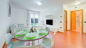 A dining area with a table set for four, a sofa, a TV, and a window with curtains. - Casa Karin With Large Terrace and Garden (Riva del Garda)
