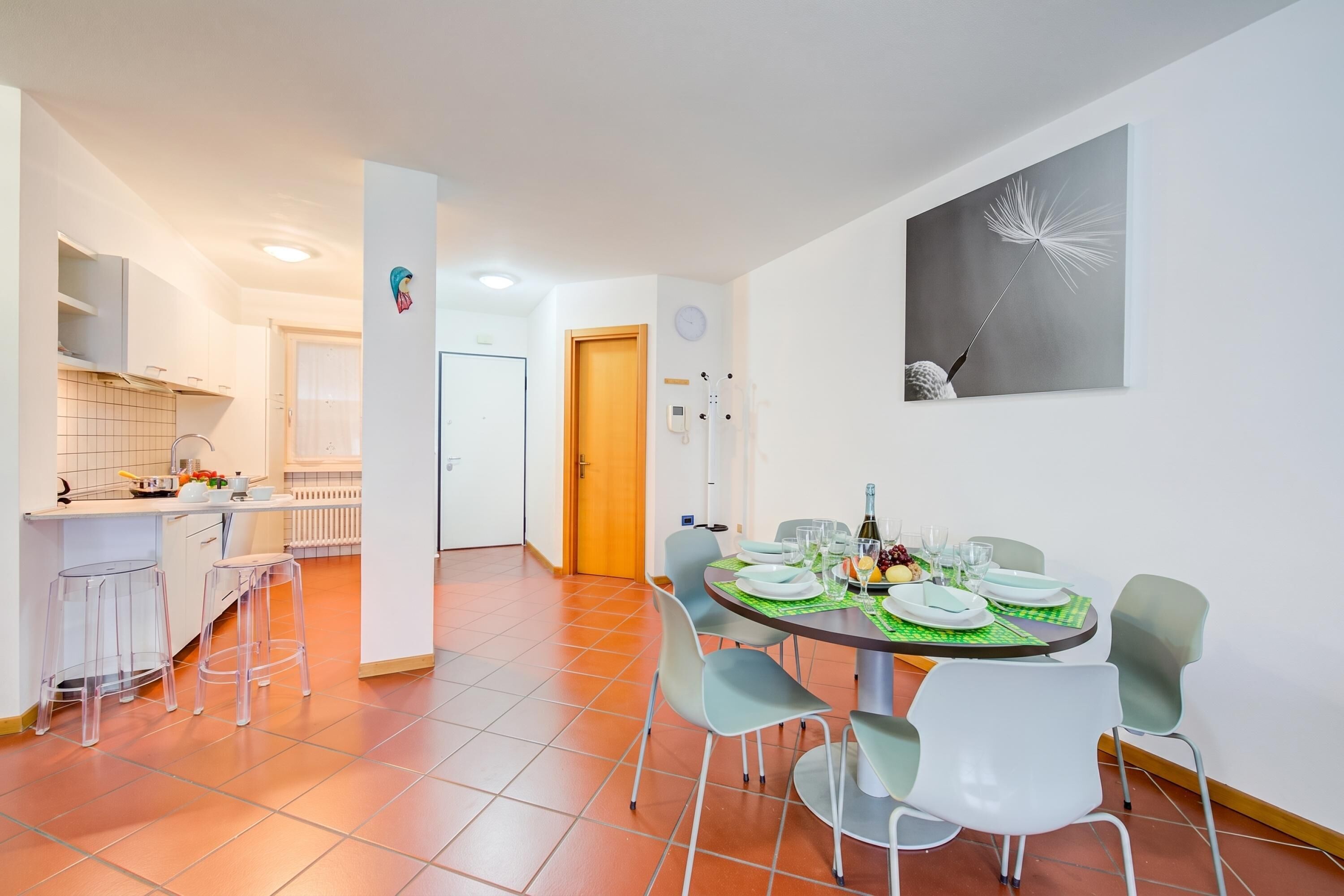 Una cocina moderna con armarios blancos, un comedor con una mesa redonda dispuesta para cuatro personas y un cuadro de diente de león en la pared.