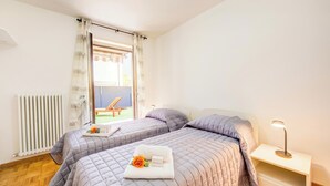 A hotel room with two beds, a radiator, a window with curtains, a nightstand with a lamp, and a view of the outside. - Casa Karin With Large Terrace and Garden (Riva del Garda)