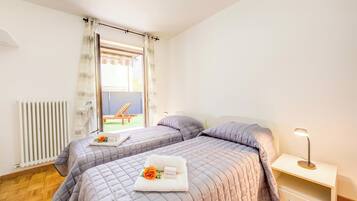 A hotel room with two beds, a radiator, a window with curtains, a nightstand with a lamp, and a view of the outside.