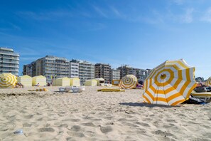 Uma praia com guarda-sóis listrados em amarelo e branco, areia dourada e edifícios ao fundo.