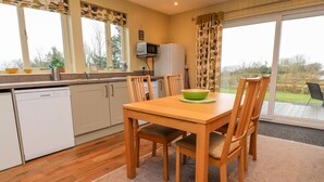 A modern kitchen with wooden cabinets, a dining table with chairs, a green bowl, and a view of the outdoors through large windows. - Oak Lodge (Preston)