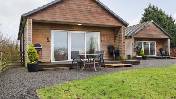 A wooden lodge with a patio area, including a table and chairs, and a small garden with a potted plant. - Oak Lodge (Preston)