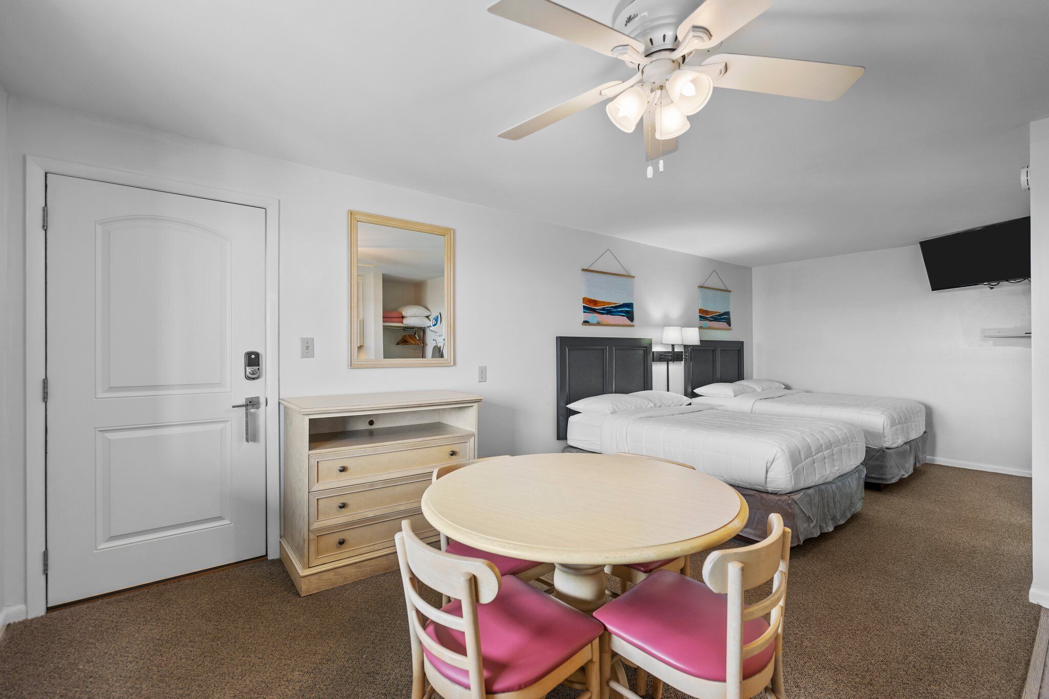 A hotel room with a white door, a desk with drawers, a round table with chairs, a bed with two pillows, a ceiling fan, and a TV mounted on the wall.