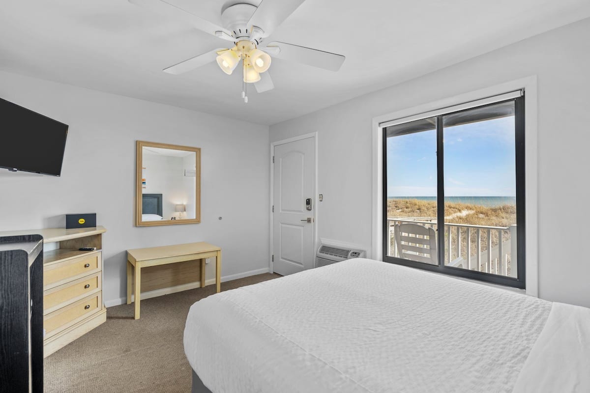A bedroom with a bed, a desk, a chair, a mirror, a ceiling fan, and a large window with a beach view.