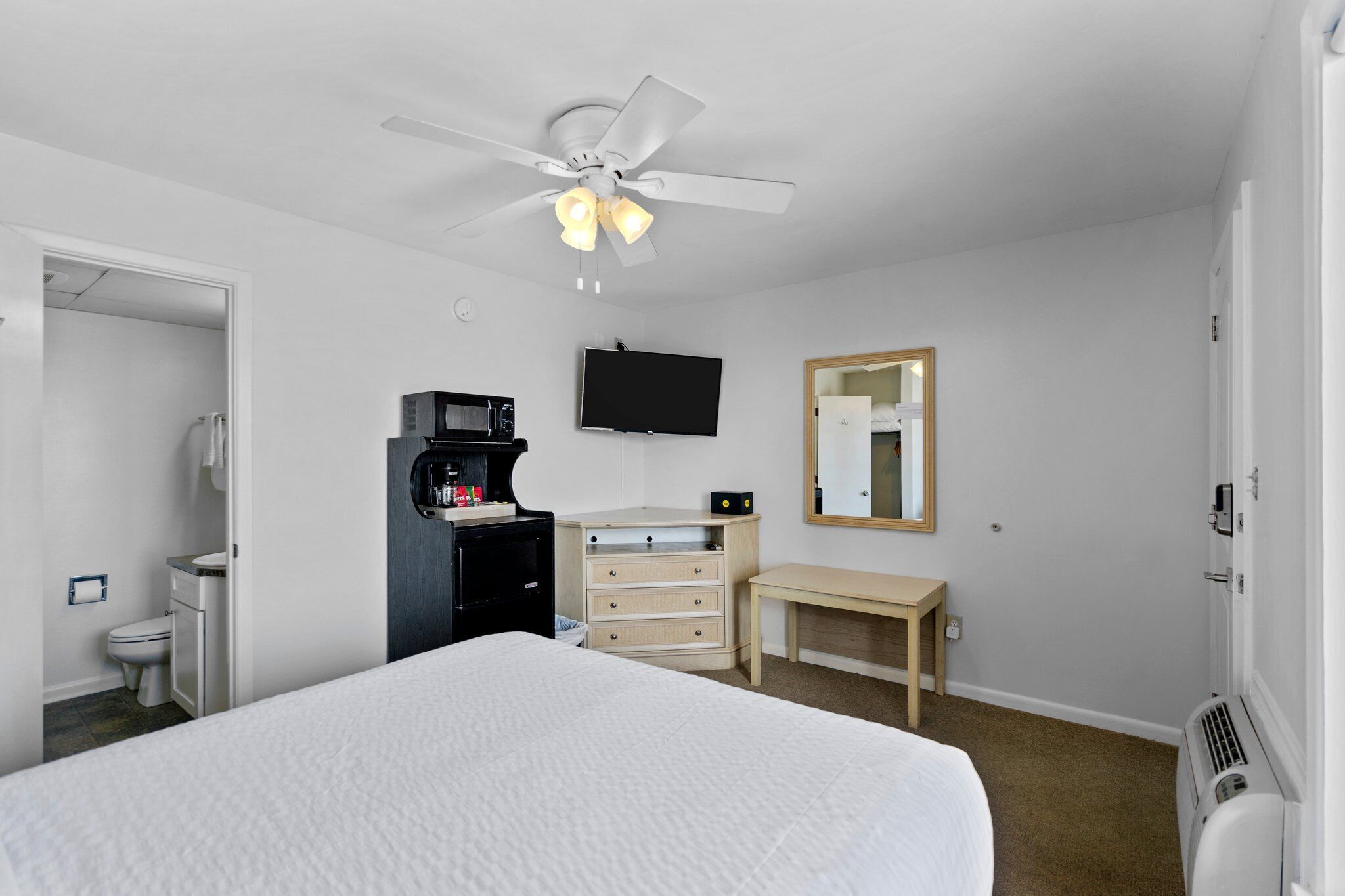 A hotel room with a bed, a desk, a chair, a television, a mirror, a ceiling fan, and a heater.