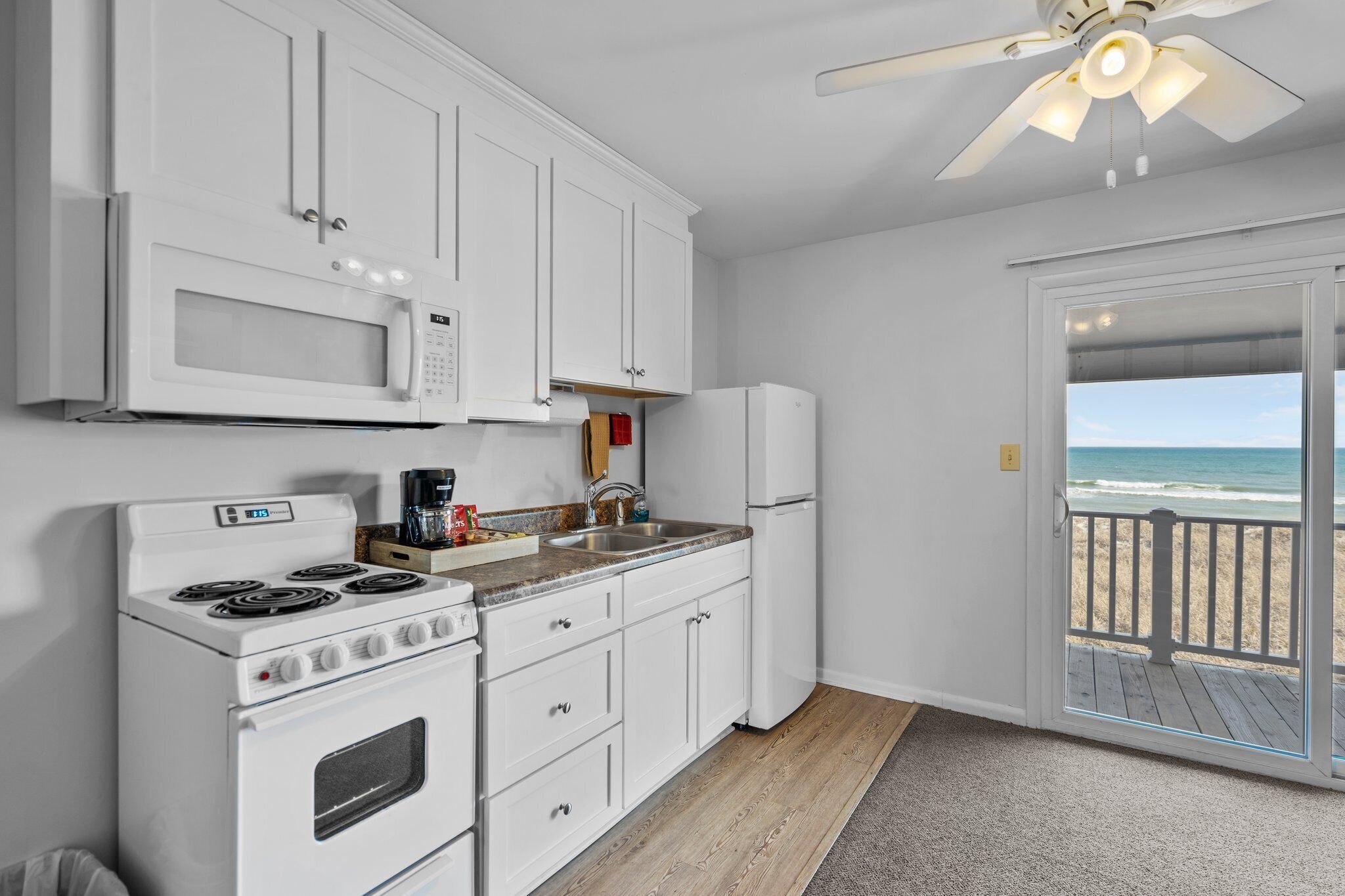 A kitchen with white cabinets, a microwave, stove, and refrigerator. There is a wooden floor and a view of the beach through a glass door.