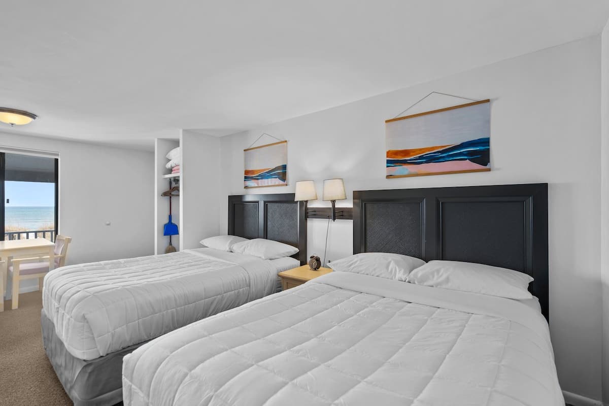 A hotel room with two beds, a desk, and a chair. There are abstract paintings on the wall and a view of the sea through the window.