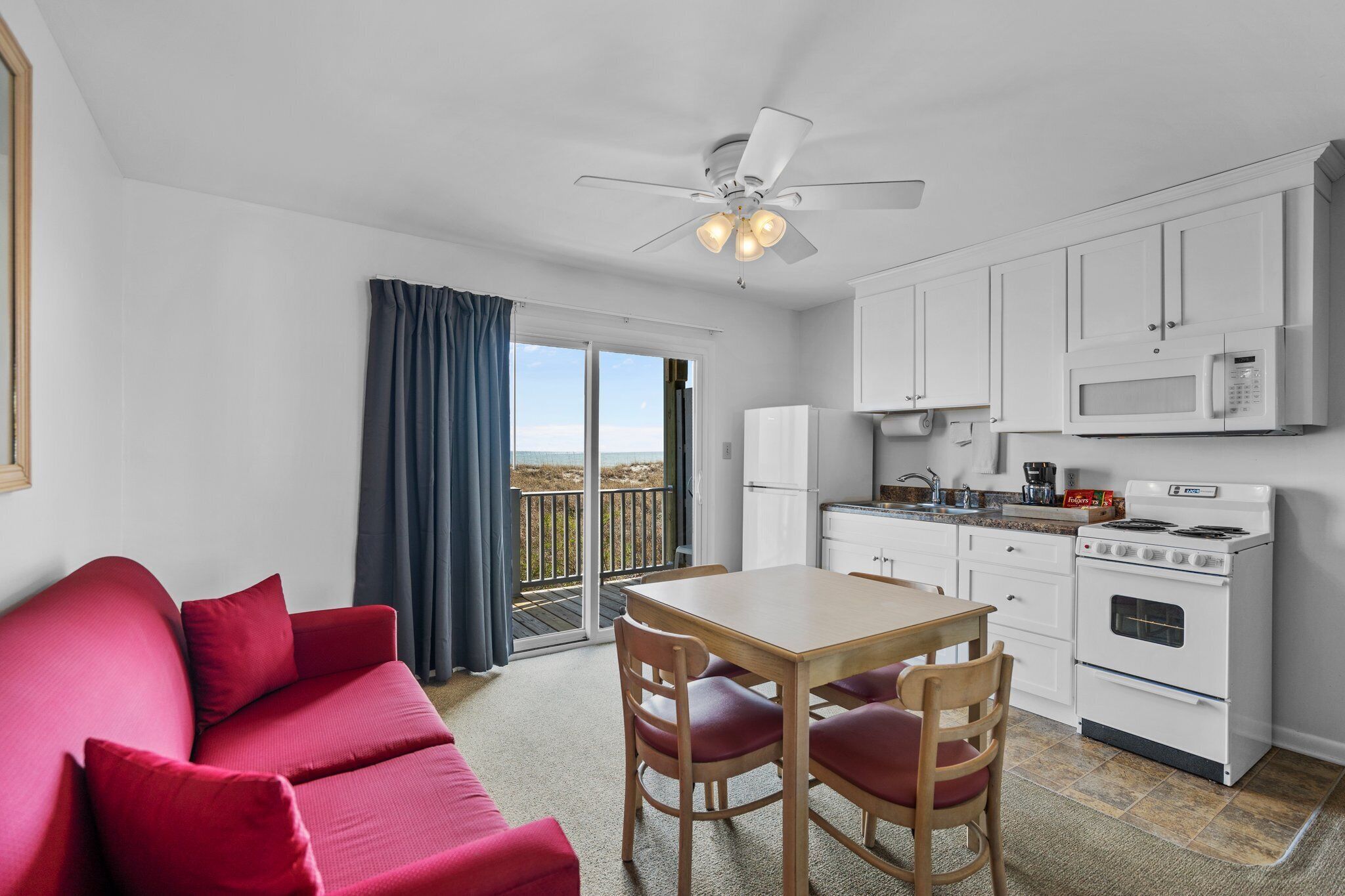 A room with a red sofa, wooden table, chairs, a white kitchen with a stove, microwave, and cabinets.