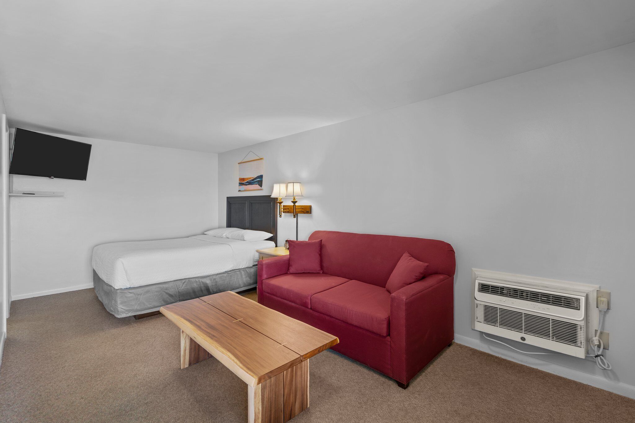 A small, modern room with a bed, a red sofa, a wooden table, a lamp, and a wall-mounted air conditioner.