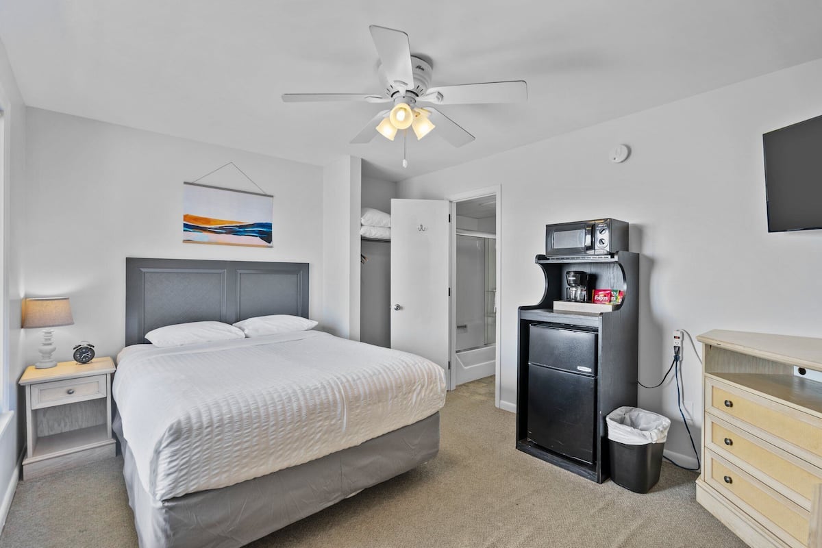 A hotel room with a bed, a nightstand, a desk, a trash bin, a microwave, a TV, and a ceiling fan.