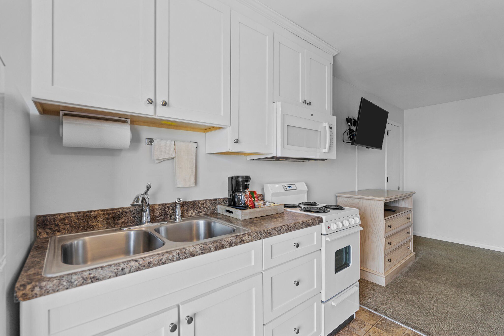A modern kitchen with white cabinets, a granite countertop, a double sink, a microwave, a stove, and a flat-screen TV mounted on the wall.