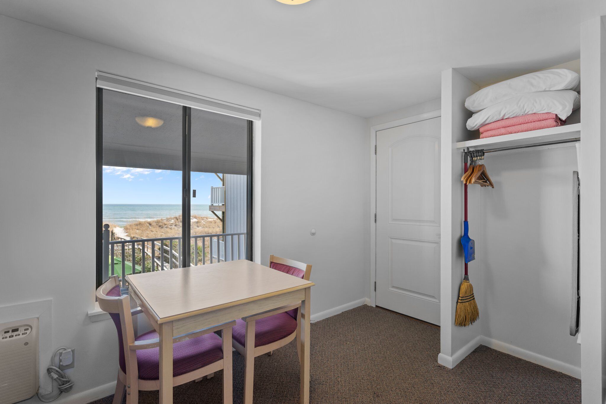 Una habitación con vista a la playa, una mesa con sillas, un armario con ropa doblada y una escoba apoyada en la pared.
