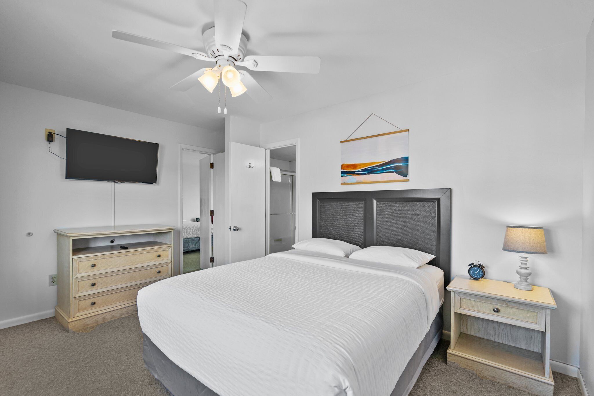 A bedroom with a bed, a nightstand with a lamp, a dresser, a ceiling fan, a TV mounted on the wall, and a painting.