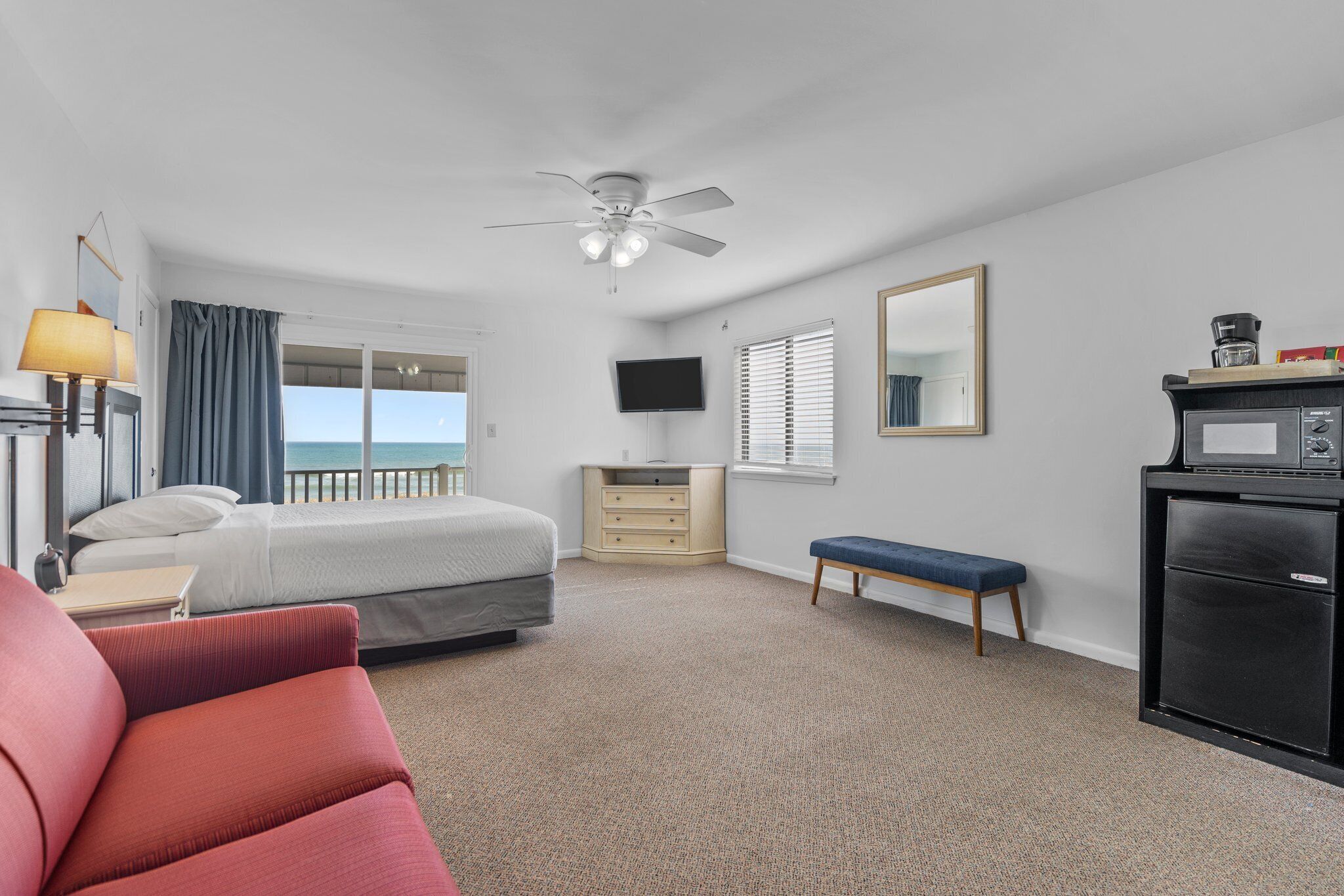 A hotel room with a bed, a desk, a chair, a bench, a television, a ceiling fan, a window with blinds, and a minibar.