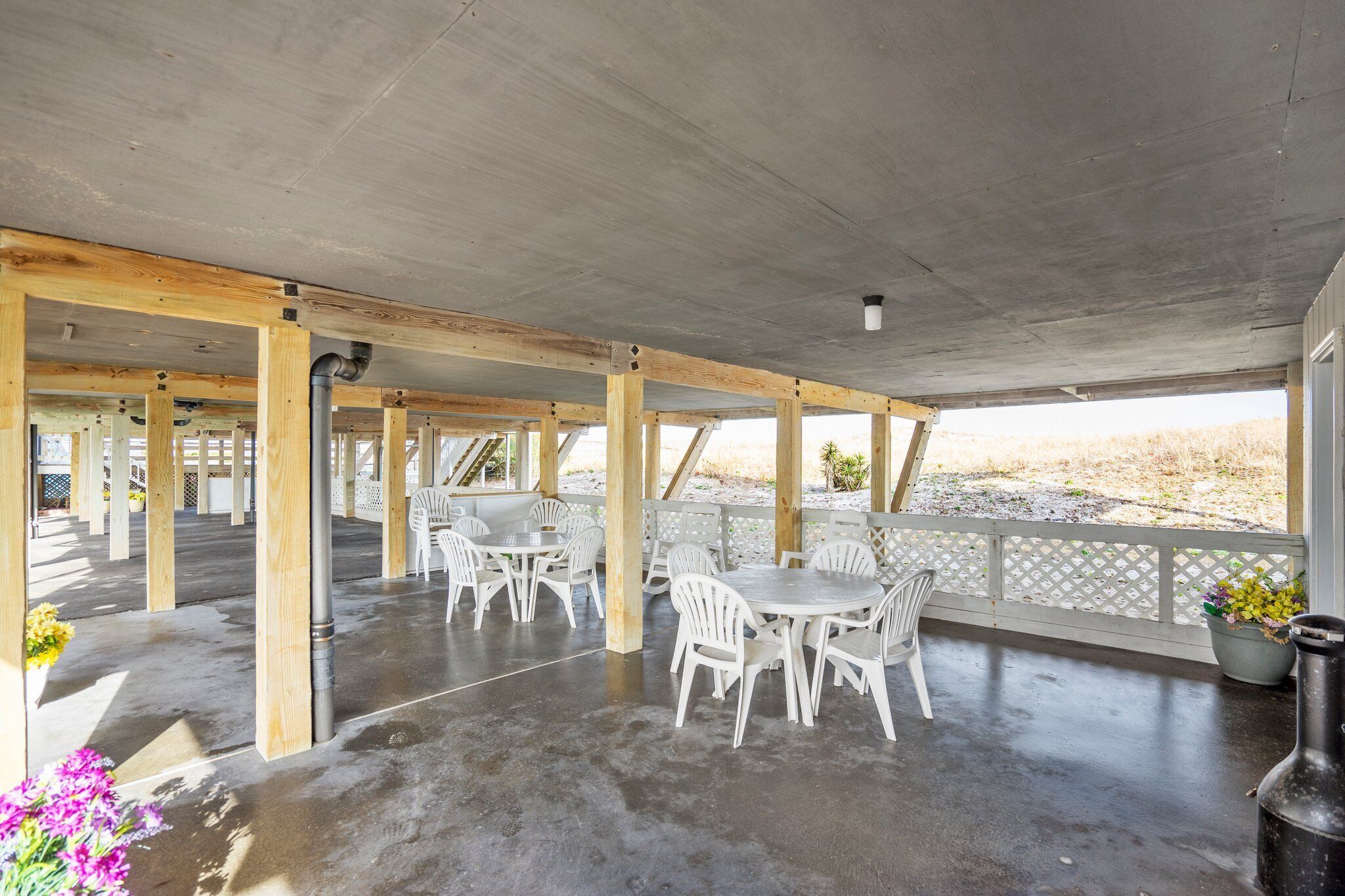 An outdoor covered patio area with concrete flooring, wooden beams, and white plastic chairs and tables.
