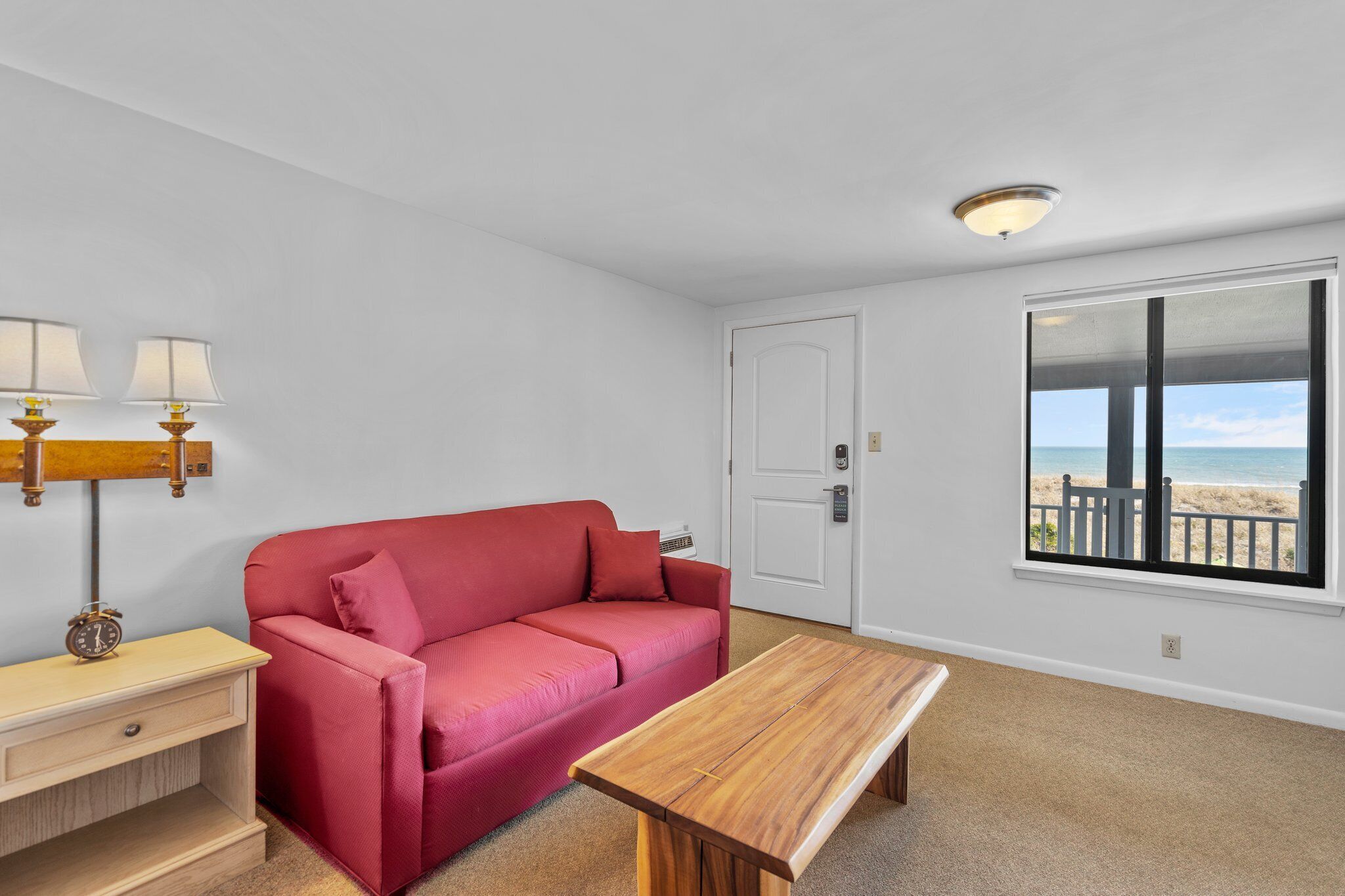 Een kamer met een rode bank, een houten tafel, een lamp en een raam met uitzicht op het strand.